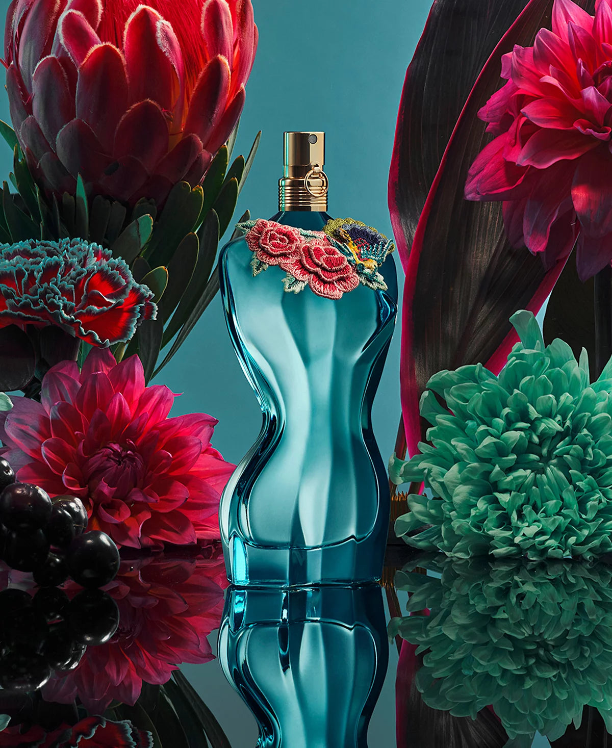 Blue perfume bottle with floral design surrounded by vibrant flowers on a reflective surface.