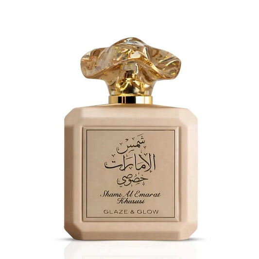 A beige rectangular perfume bottle with a sculptural gold cap, featuring Arabic calligraphy and the text 'Shame Al Emarat Khususi' and 'GLAZE & GLOW' on the front label.